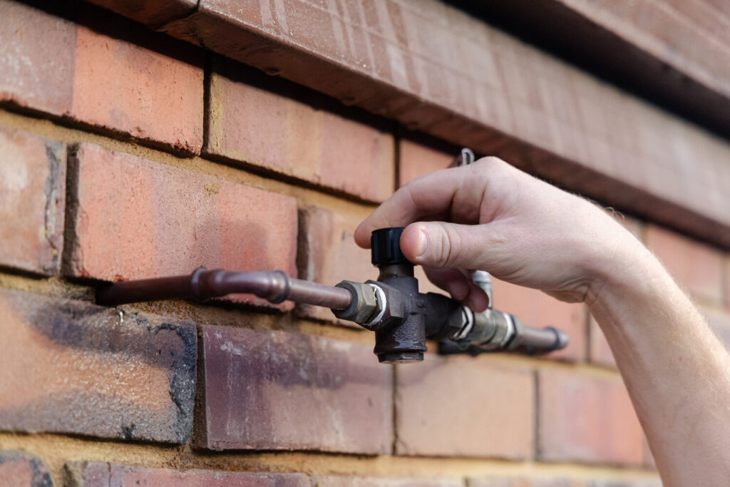 A hand turning a black valve fitted to copper pipe on a red brick wall
