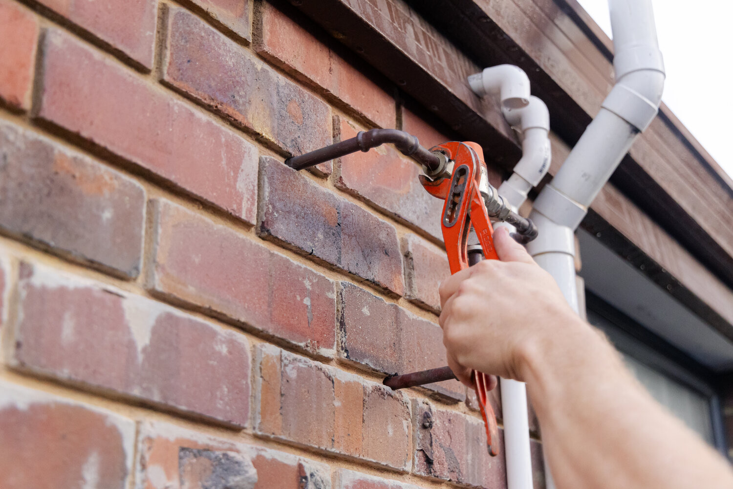 Plumber's hand tightening a copper pipe fitting with orange-handled cutters beside a white PVC downpipe
