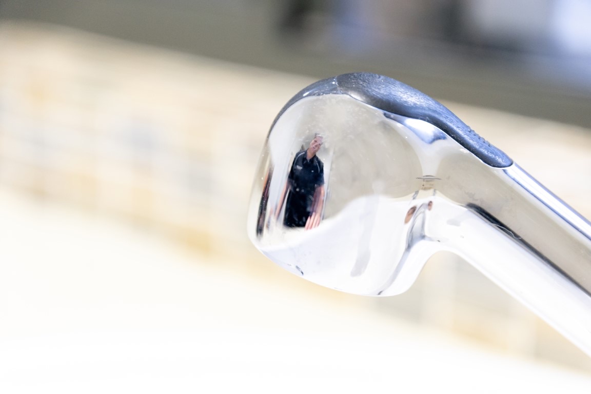 Chrome tap spout in sharp focus, a figure in dark clothing mirrored in the metal